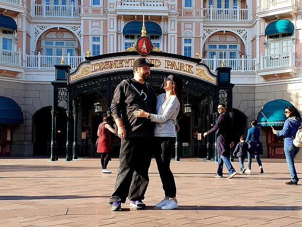 Benjamin Kott und Darina Schkolnik stehen Arm in Arm vor dem Eingang des Disneyland Parks in Paris und lächeln sich an.